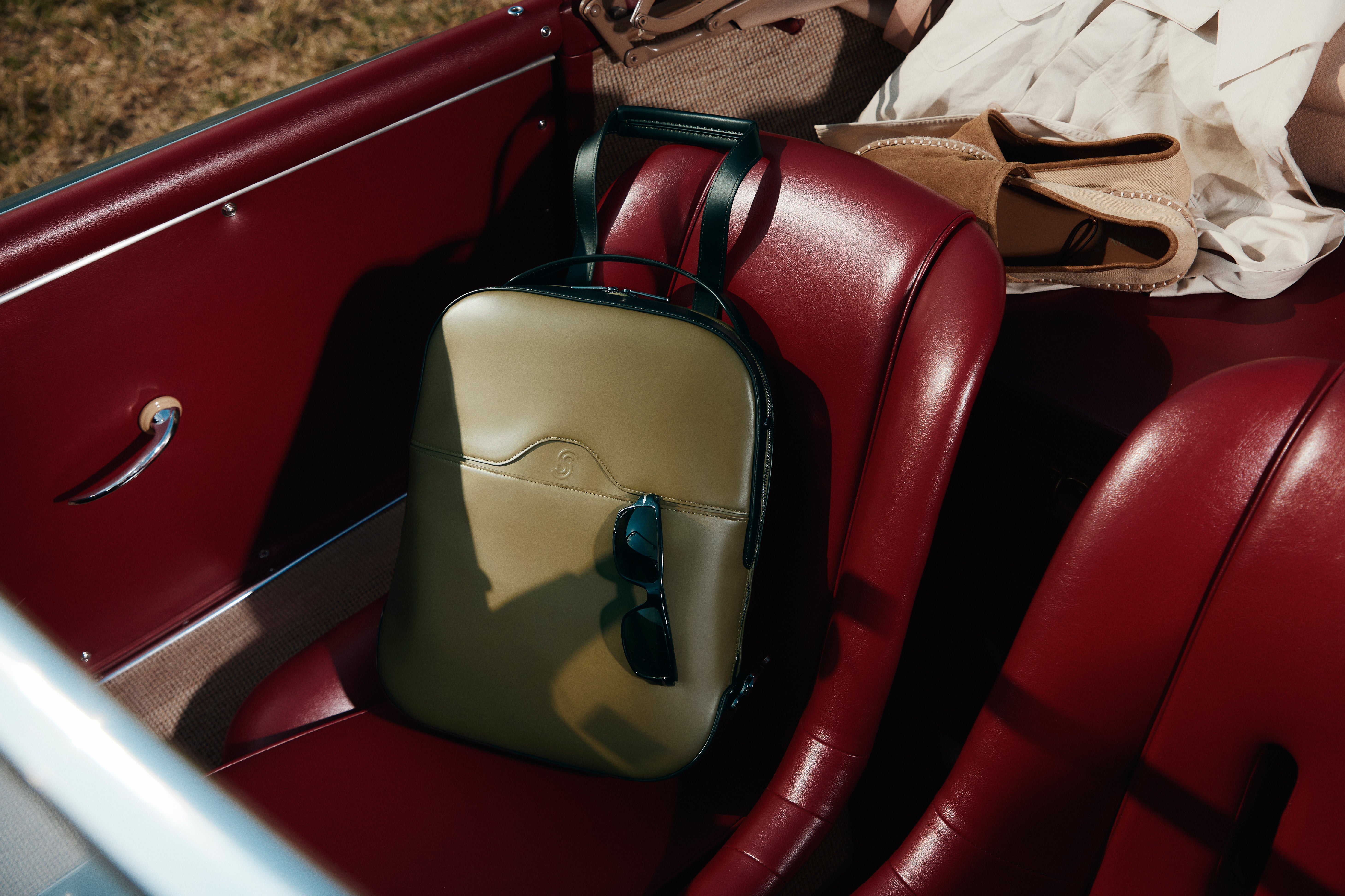 A structured olive green leather backpack with black handles and a small shield logo on the front, resting on a red leather car seat with sunglasses hanging from its side.